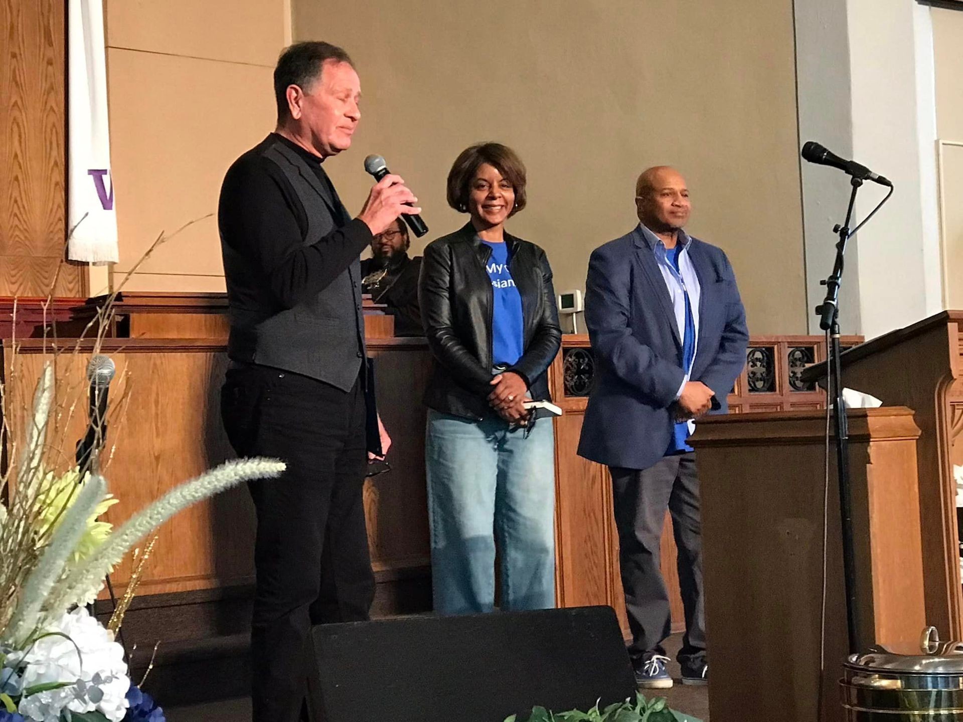 Three people standing on a stage with microphones in a church setting, addressing the audience.