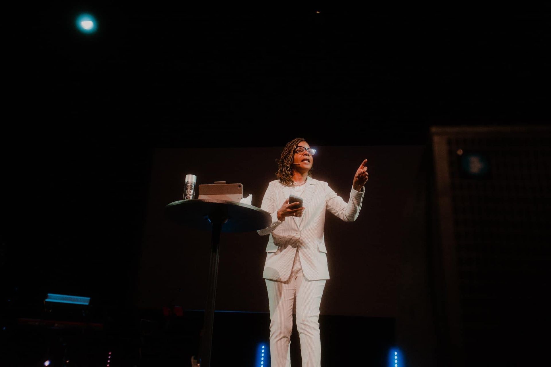 Speaker in white suit delivering a presentation on stage, holding a phone and facing the audience.